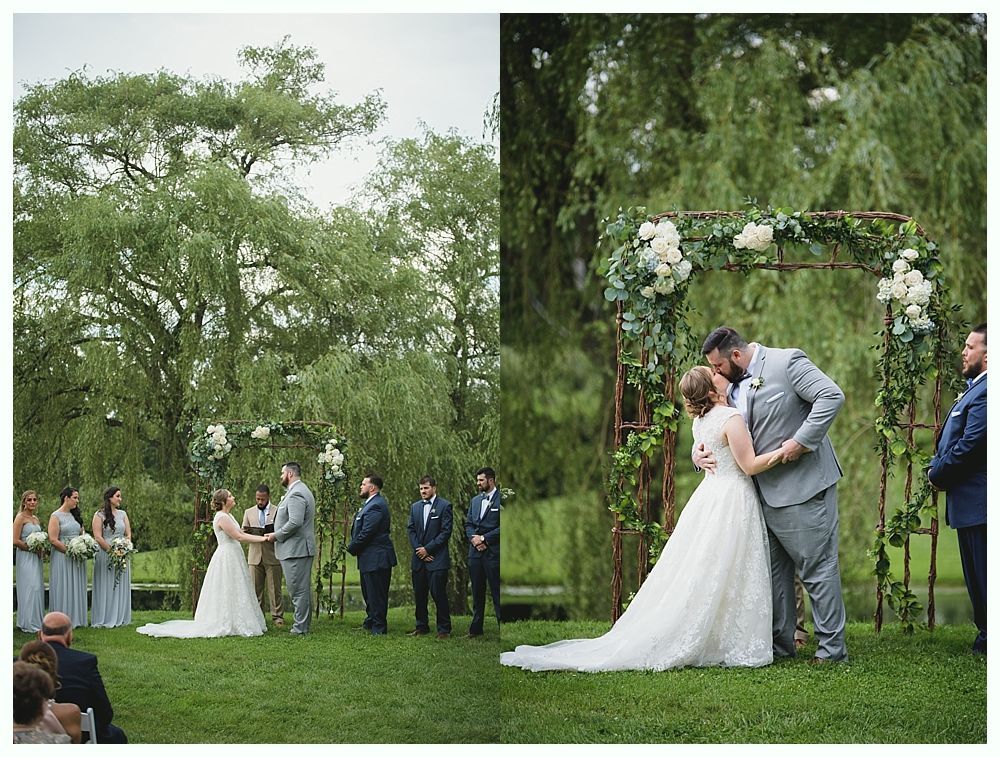 Wedding ceremony: Couple kissing under floral arch, surrounded by wedding party on green lawn, trees in background.