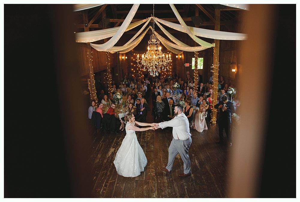 Bride and groom dance at reception in a rustic barn with guests watching and a chandelier.