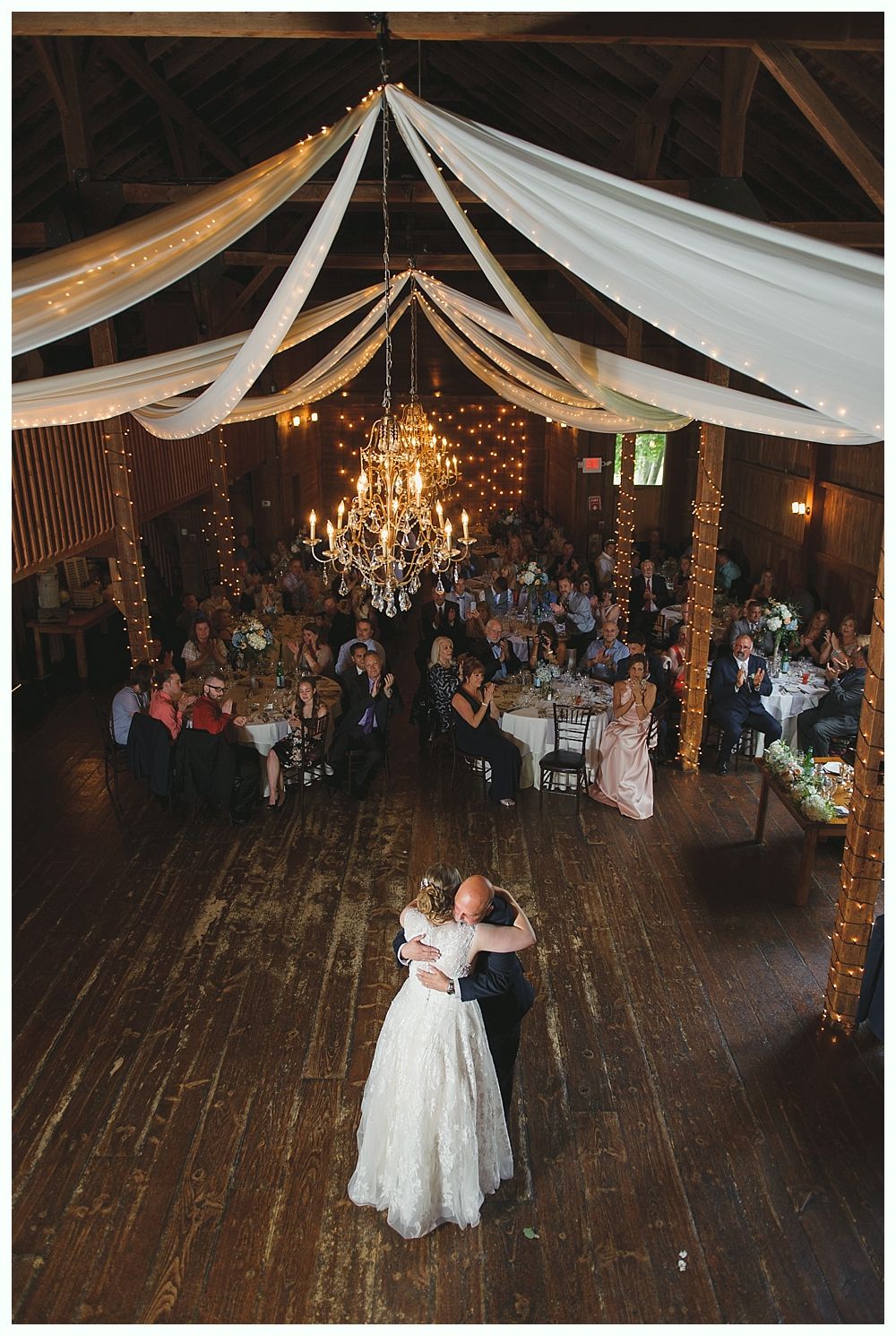 Bride and groom dance in a decorated barn. Guests seated at tables watch. Fairy lights and a chandelier hang.