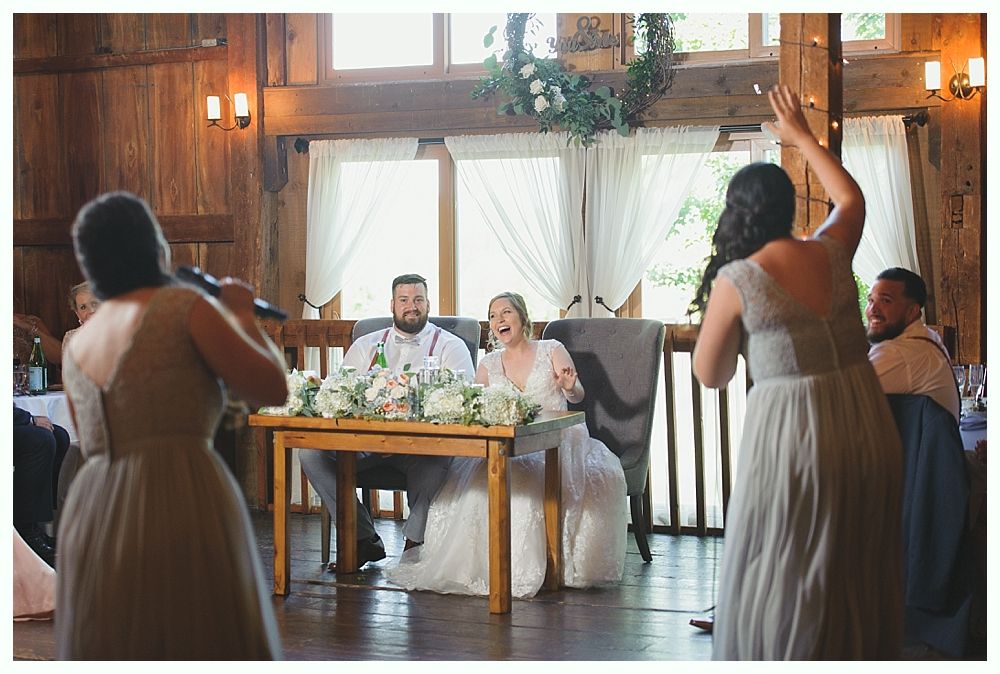 Wedding reception: Bride and groom at table, guests celebrating with cheers and applause. Wooden interior, light colors.
