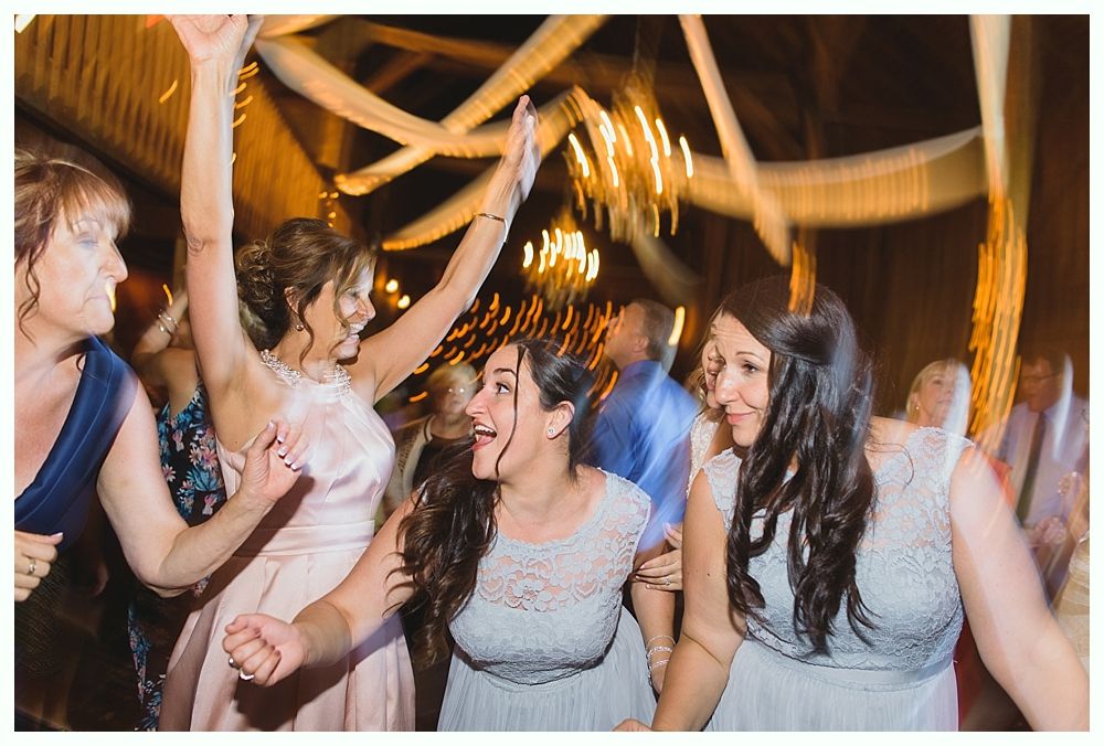 People dancing at a celebration, indoors. Some have raised arms, joyful expressions. Warm lighting, blurred background.