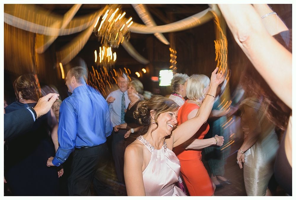 People dancing at an event, blurry movement. Woman in pink dress smiles, raising arm. Yellow lights and fabric.