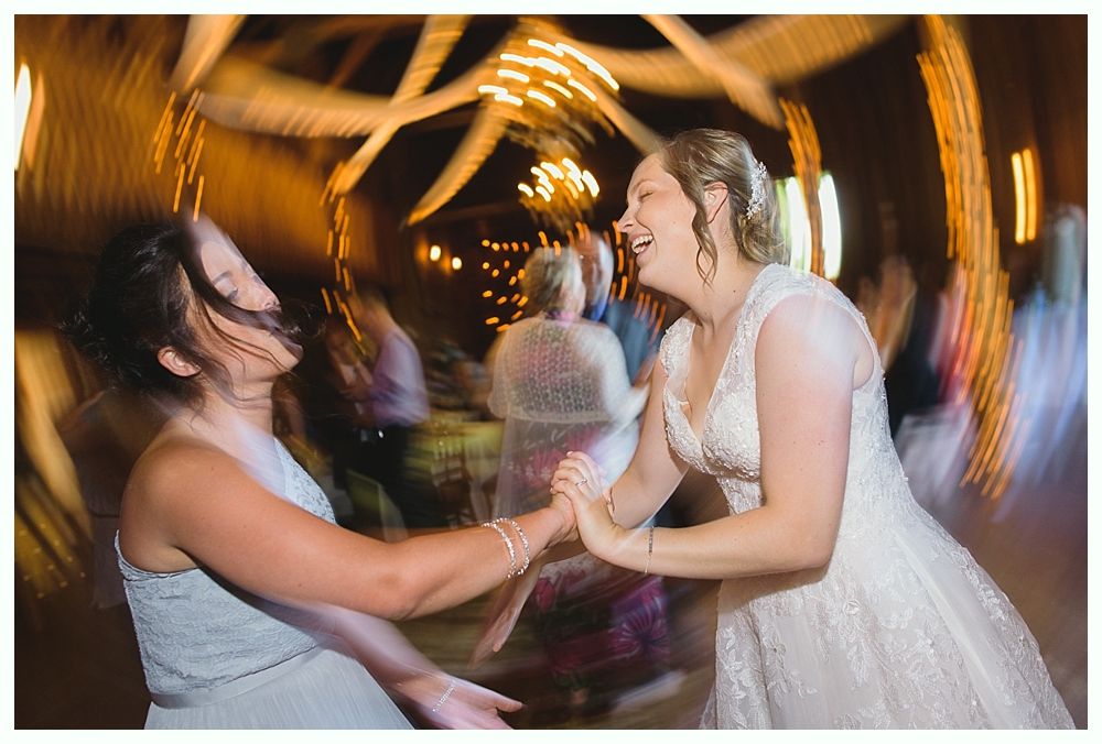 Bride and friend laughing, dancing at a wedding reception. Blurred motion, warm lighting, celebratory atmosphere.