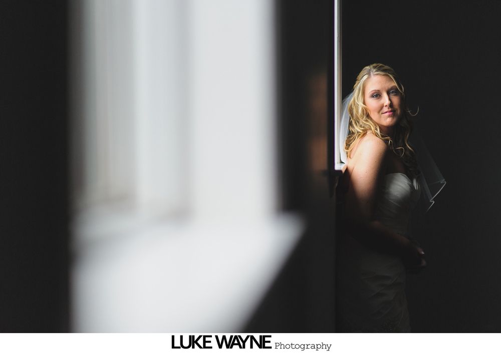 Bride in wedding dress, leaning in doorway, lit by sunlight.
