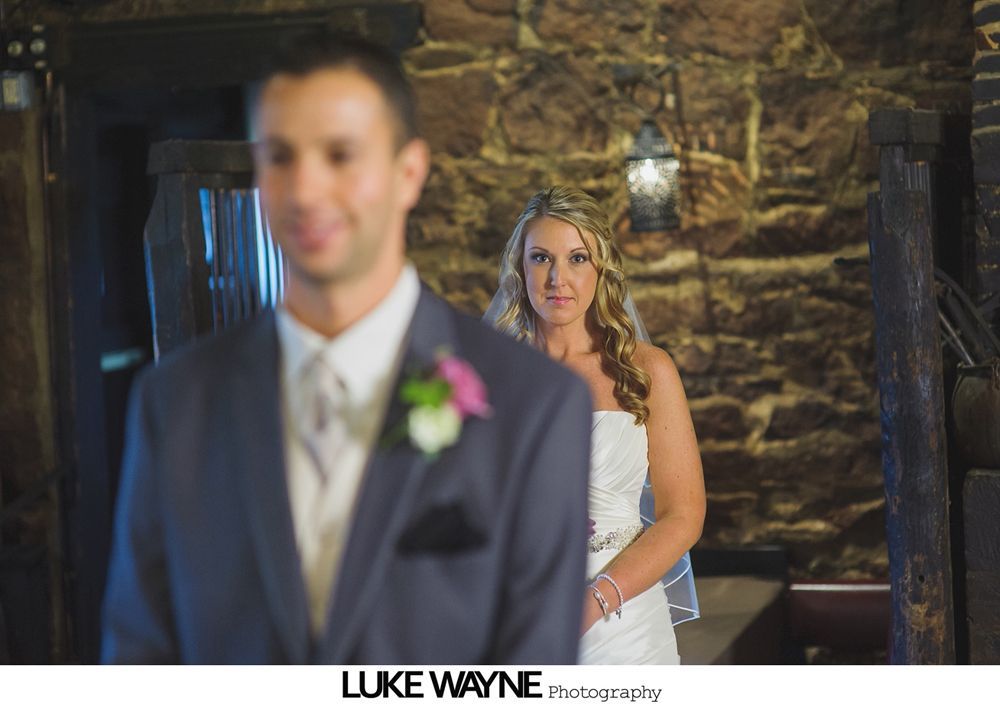 Bride approaches groom from behind; both are in formal wedding attire. Stone walls in the background.