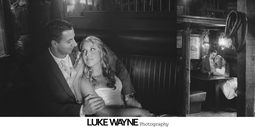 Bride and groom embrace in a booth. Another person sits at a table in the background. Black and white photo.