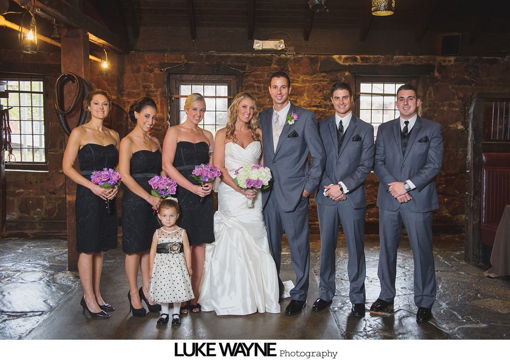 Wedding party posing indoors, bride in white dress, bridesmaids in black, groomsmen in gray.