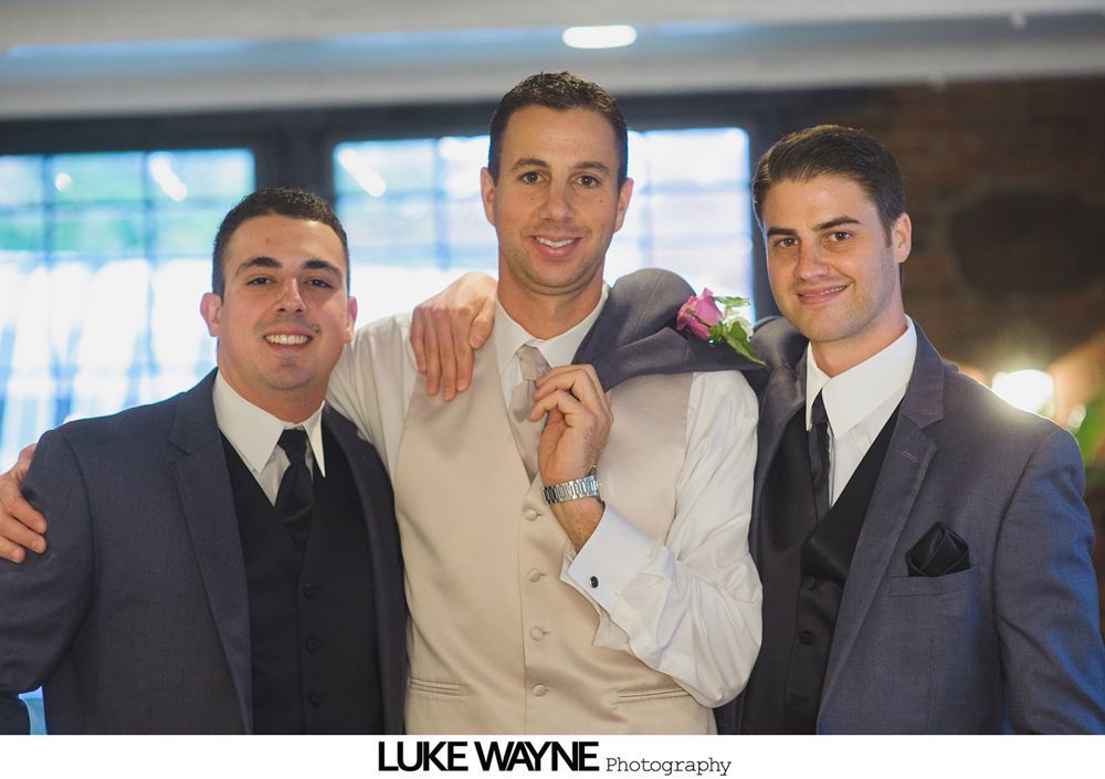 Three men in suits pose indoors, the center man holding a suit jacket.