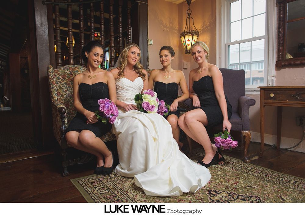 Bride and bridesmaids in black dresses seated, posing, holding bouquets. Ornate interior, antique furniture.