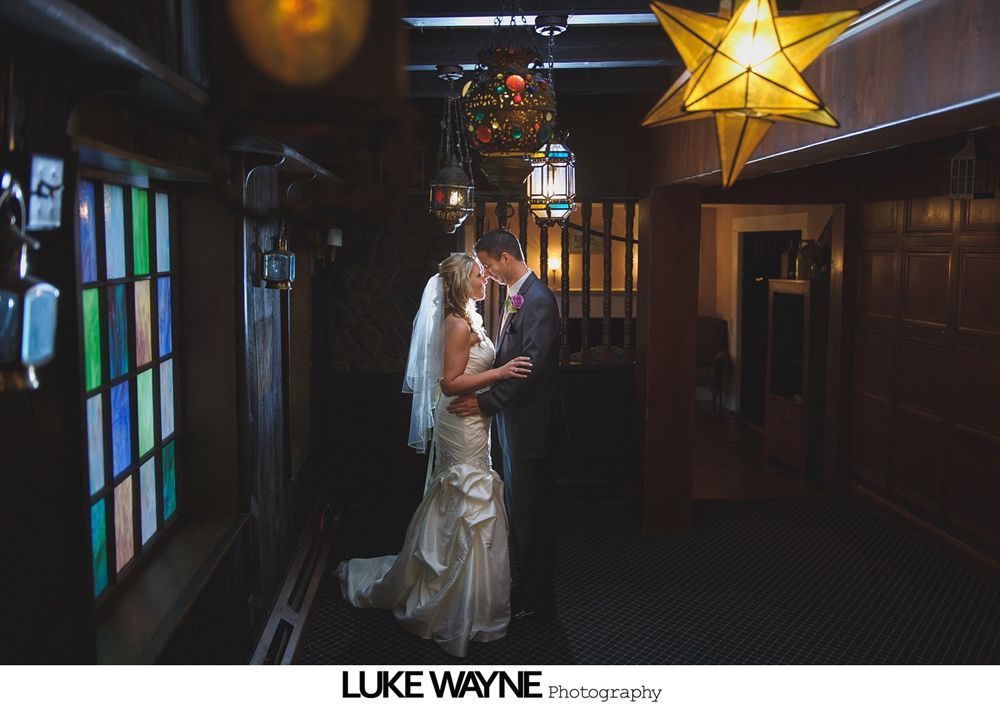Bride and groom embrace in a dimly lit hallway; stained glass window, star lantern, and hanging lights overhead.