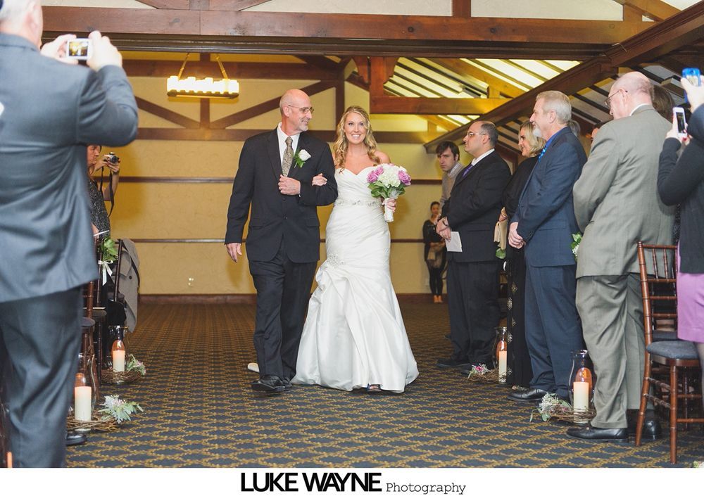 Bride in white gown walks down the aisle with her father, guests watch. Indoor wedding setting.