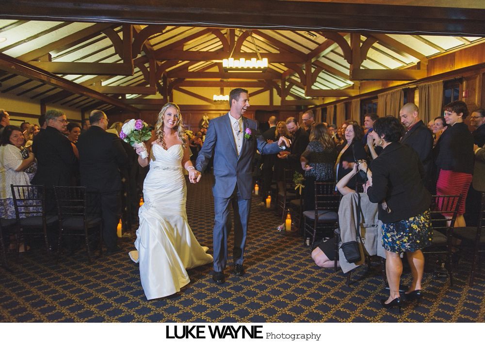 Newly married couple walks down aisle, holding hands, smiles. Indoor wedding. Guests watch and clap.