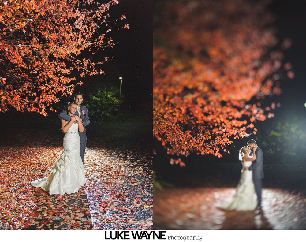 Couple in wedding attire under red-leafed tree at night, spotlit with blurred bokeh effect.