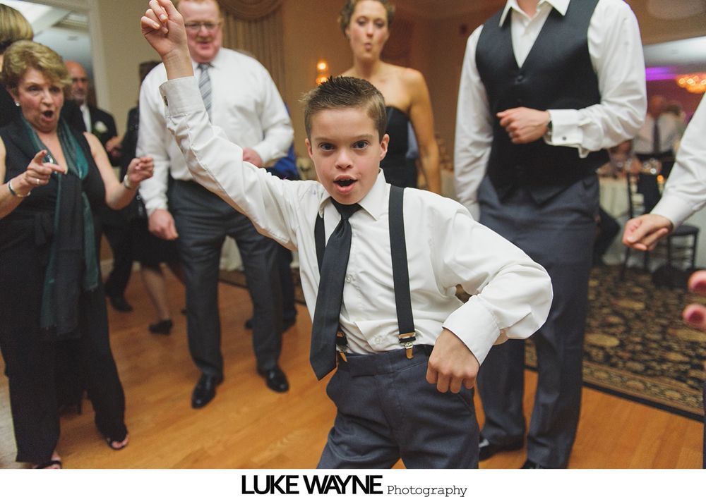Boy dancing energetically at a formal event, wearing suspenders and tie; others dancing nearby.