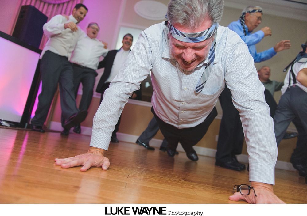 Man in business attire doing push-ups on a wooden floor, with other men cheering and laughing in a party setting.