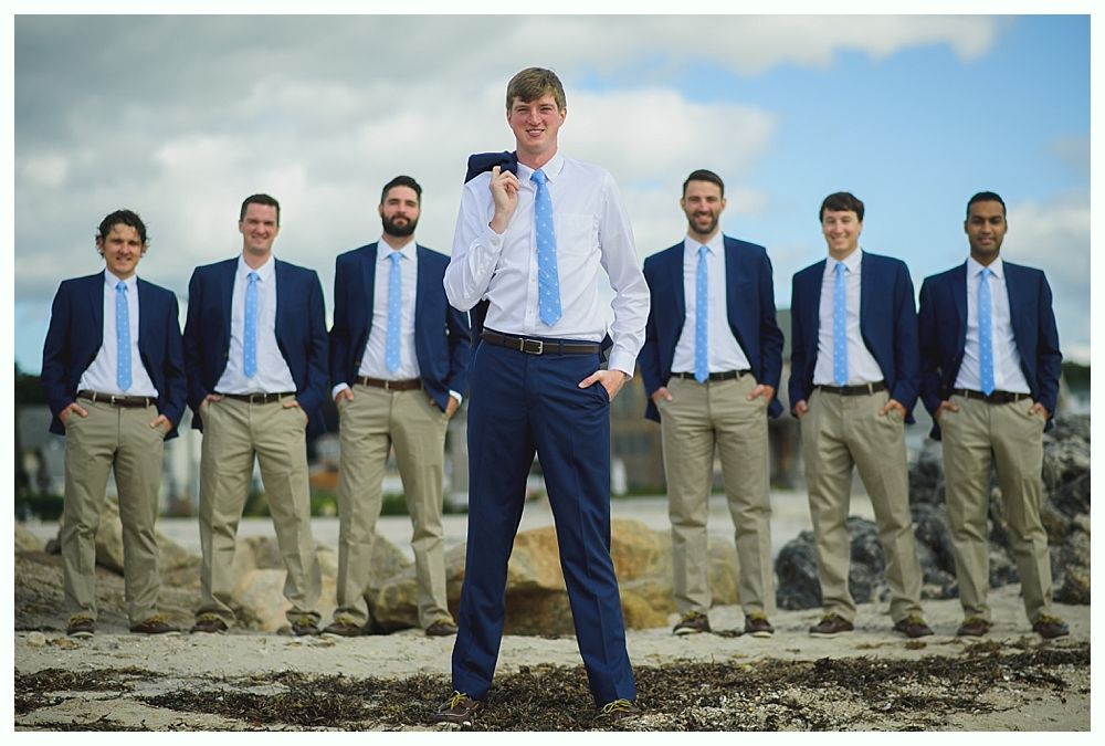 Groom and groomsmen on a beach, wearing suits. Groom in navy pants, others in khaki. All with blue ties, ocean in background.
