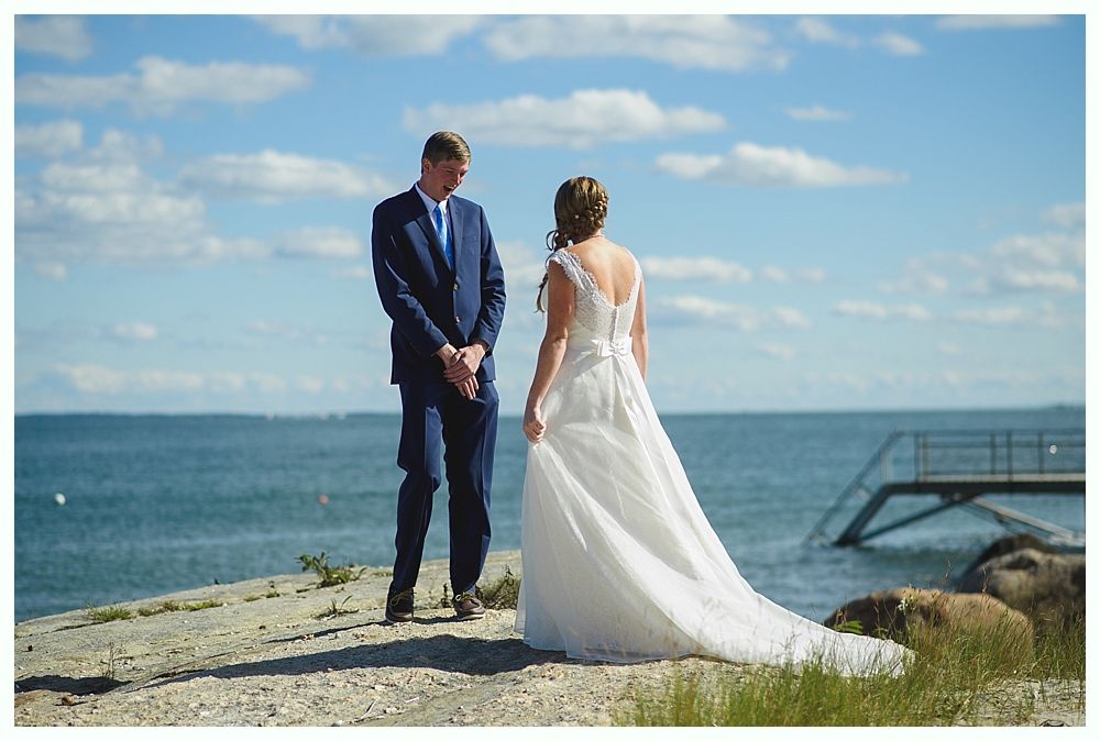 Bride and groom on rocky shoreline, ocean and blue sky background. Groom in suit, bride in white dress, looking at each other.