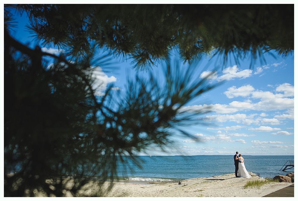 Bride and groom embrace on a beach. Ocean, blue sky, and tree branch in the foreground.