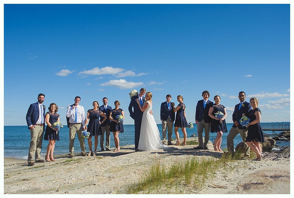Wedding party on a beach with blue sky. Bride and groom embrace, others stand nearby. Dark blue dresses and jackets.