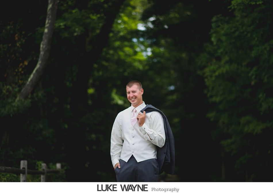 Man in vest and suit jacket smiles, holding jacket over his shoulder, standing in a tree-lined path.