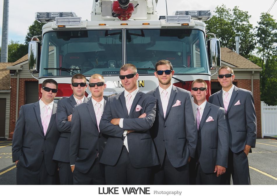 Groomsmen in gray suits and sunglasses pose in front of a fire truck, with pink pocket squares.