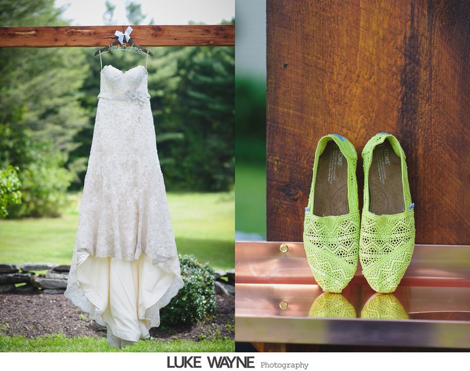 Wedding dress hanging on a wooden beam outdoors, next to green lace shoes on a wooden surface.