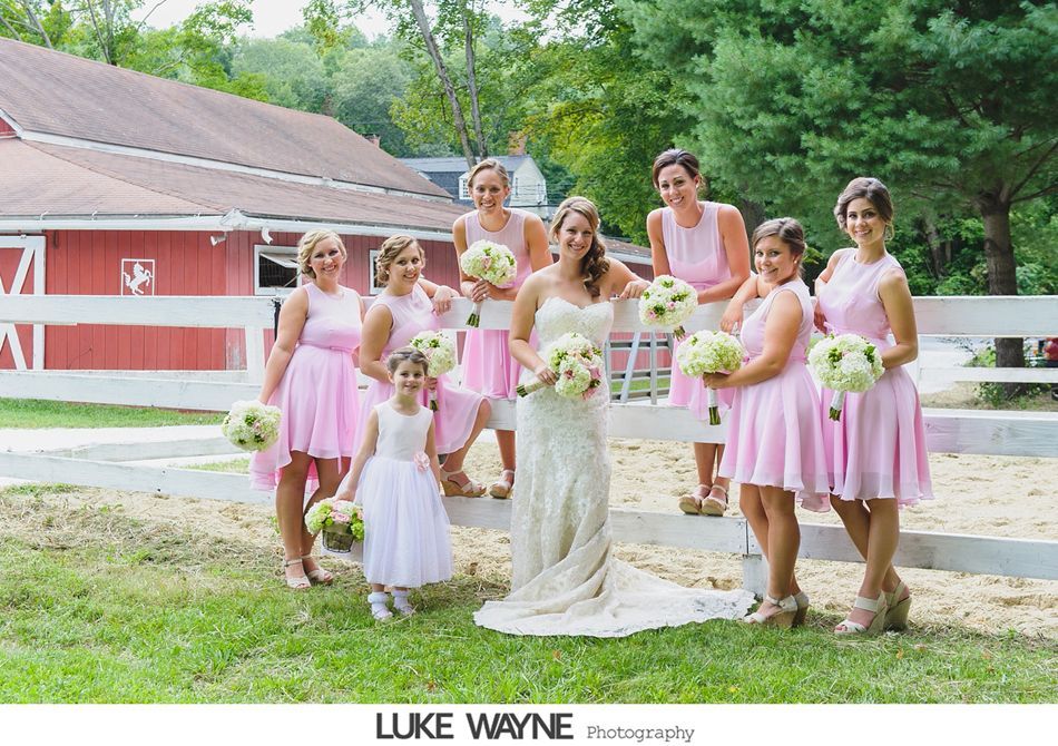 Bride and bridesmaids in pink dresses pose by a white fence, red barn background.