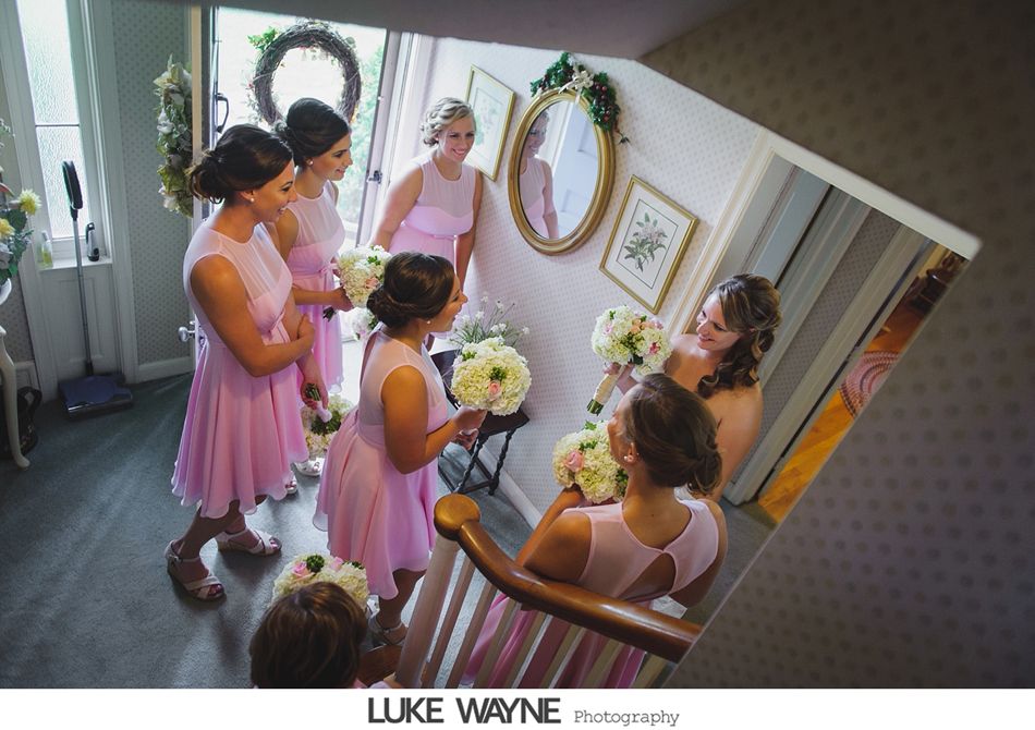 Bridesmaids in pink dresses hold bouquets on a staircase. One looks in a mirror with flowers.