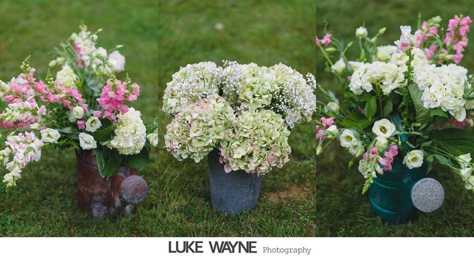 Three floral arrangements in boots, a pot, and a watering can on grass.