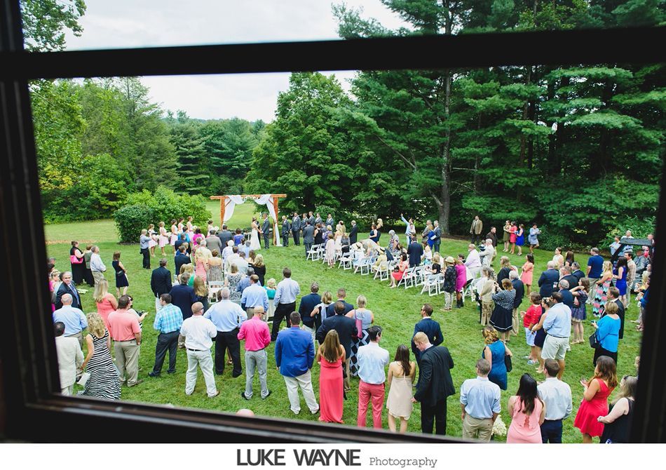 Wedding ceremony in a grassy outdoor setting, viewed through a window. Guests watch as couple exchanges vows.