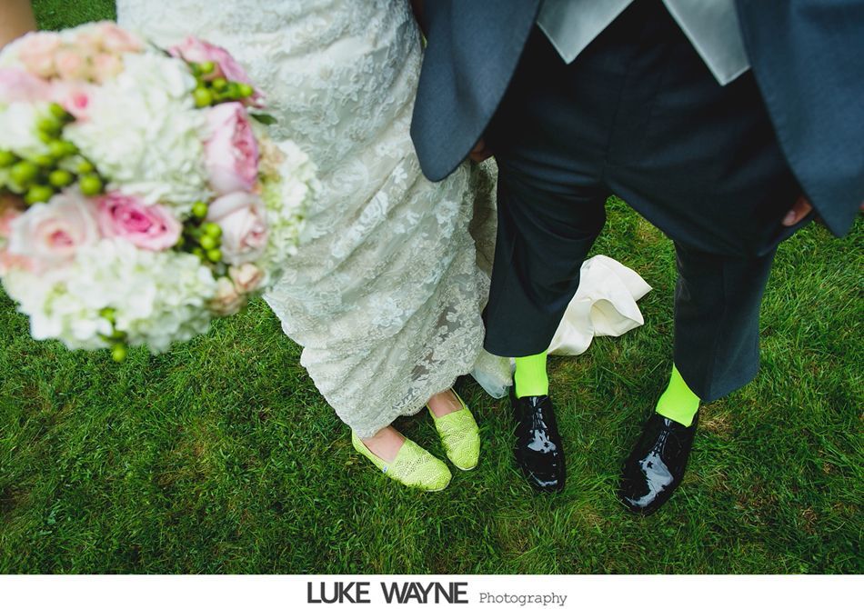 Bride and groom stand on grass; bride in lace dress, yellow shoes; groom in suit, black shoes, neon socks; holding bouquet.