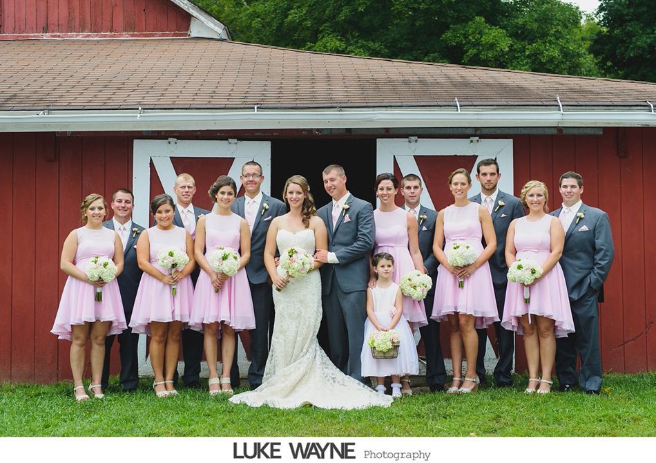 Wedding party in pink dresses and gray suits pose in front of a red barn.