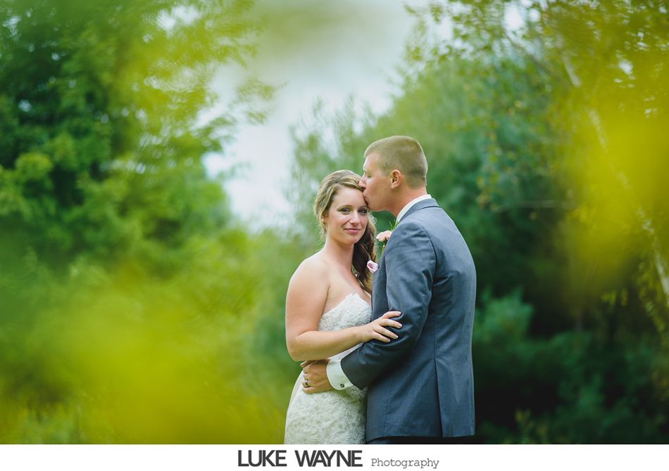 Groom kisses bride's forehead. They embrace in a green, outdoor setting, framed by blurred foliage.