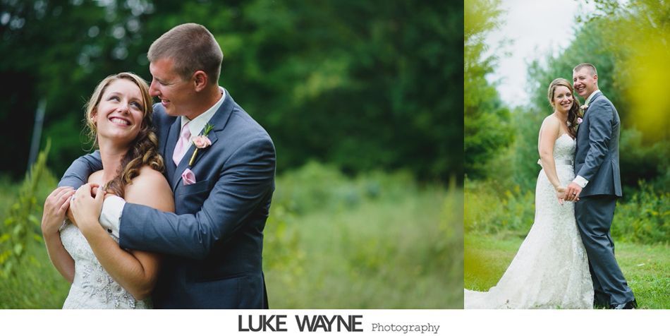 Bride and groom embrace in a field, smiling. The man wears a suit, and the woman has a white dress. Green trees in the background.
