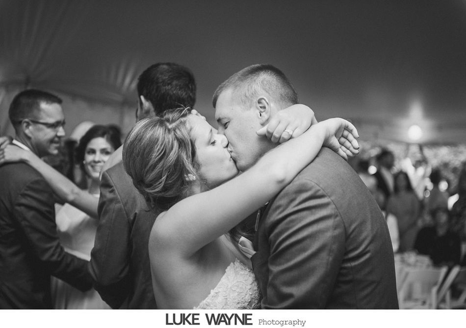 Bride and groom kiss during a wedding reception, embraced by guests. Black and white photo.