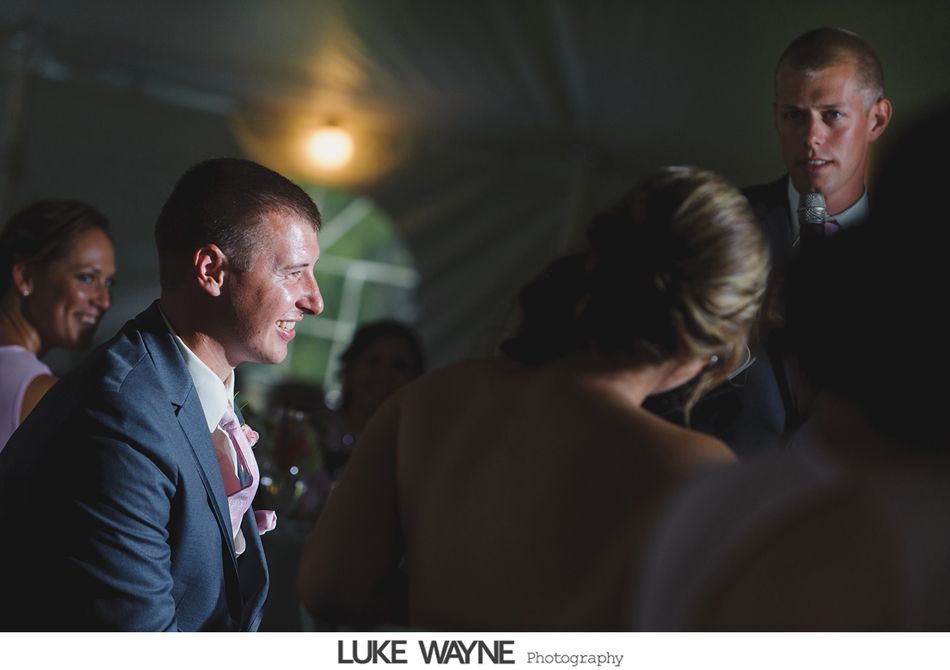 Man in blue suit smiles, talking with others at an event, overhead light, tent setting.