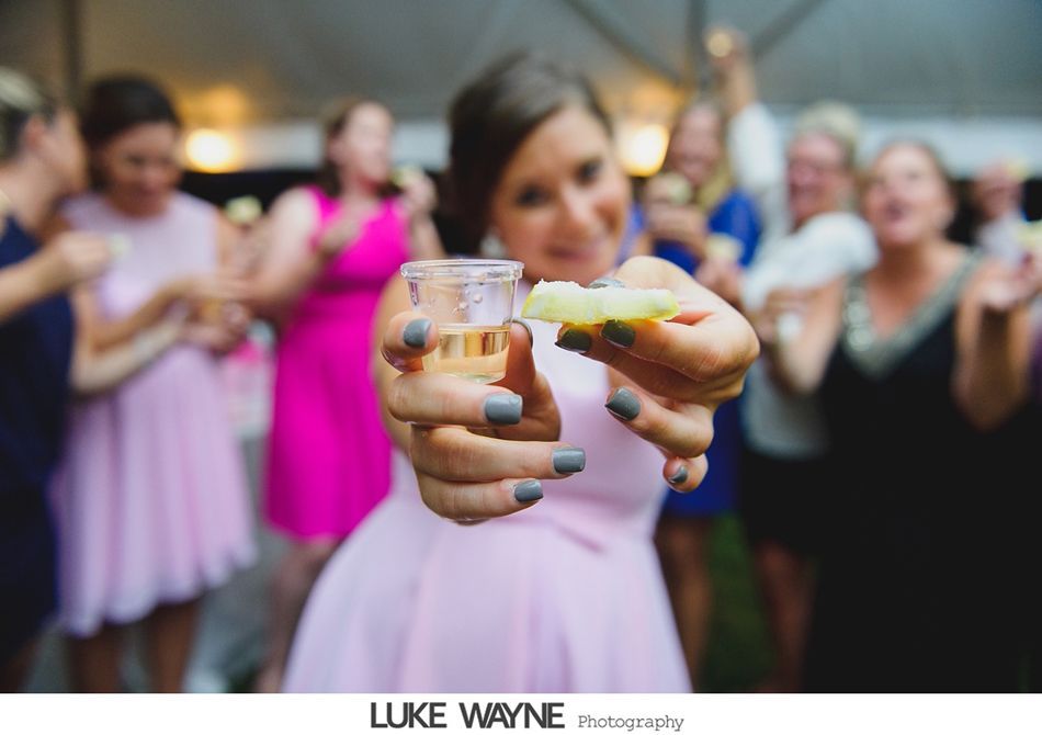 Woman in pink dress holds a shot of alcohol with lime, smiling in front of a wedding party.