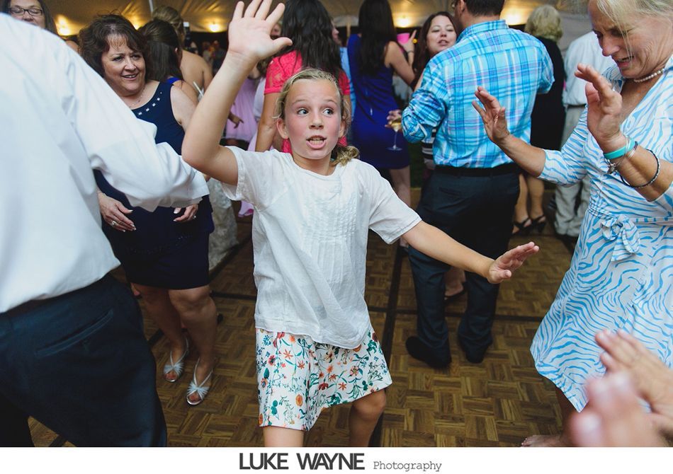 Girl enthusiastically dancing at a party, surrounded by other people dancing on a wooden floor.