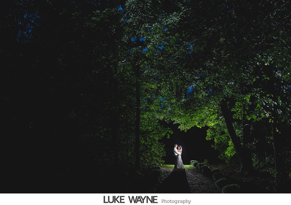 Couple embraces on a path, illuminated by green light beneath dark trees.