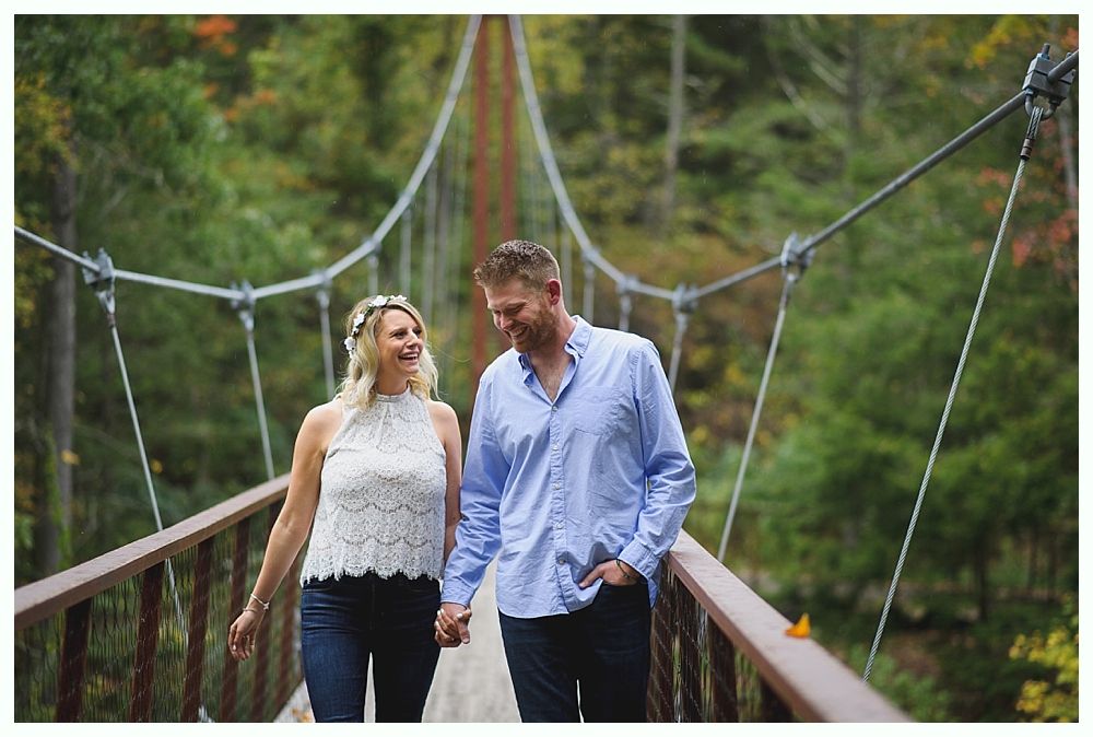 Couple holding hands, smiling while walking on a wooden suspension bridge in a forest.
