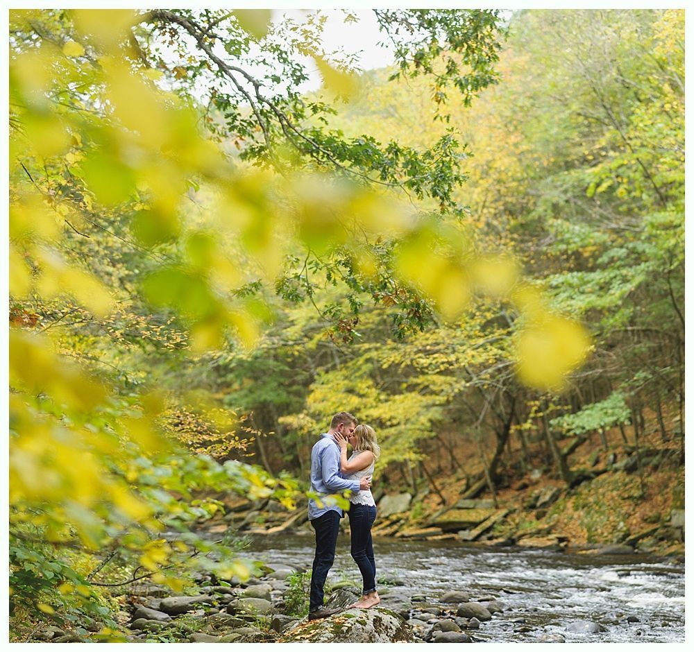 Couple kissing by a stream in autumn. Surrounded by trees with yellow and green leaves.