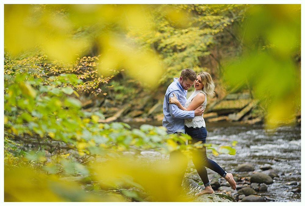 Couple embracing by a river, blurred foliage in foreground; autumn colors.