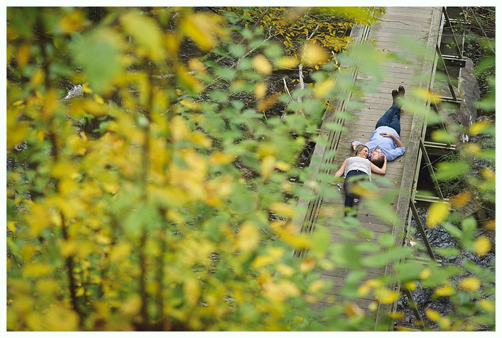 Couple lying on a wooden bridge, surrounded by fall foliage. Yellow and green leaves obscure the view.