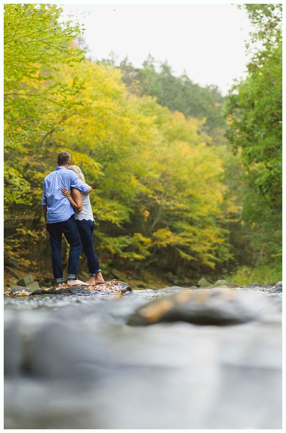 Couple standing on rocks in a creek, surrounded by colorful fall trees.