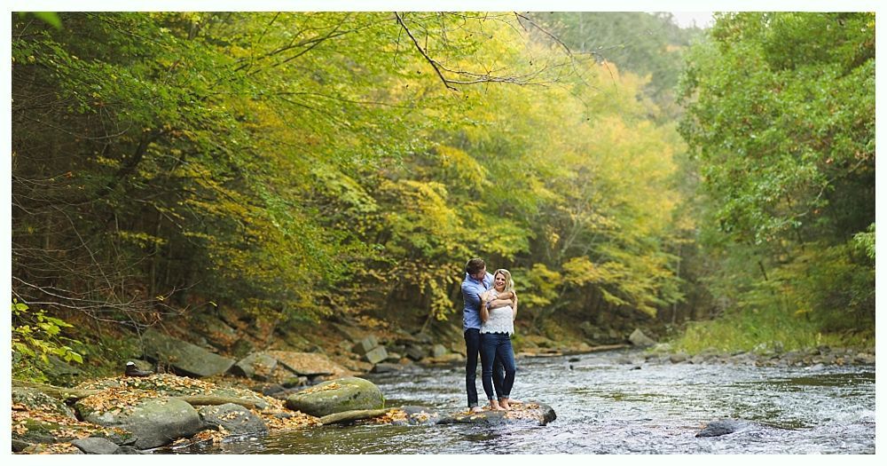 Couple standing in a shallow stream, hugging; surrounded by green trees in a sunlit forest.