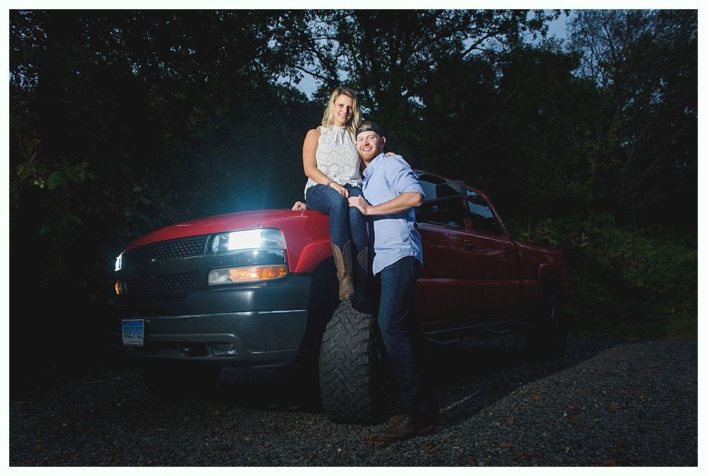 Woman sitting on red truck hood; man standing by tire, smiling. Nighttime setting, headlights on.