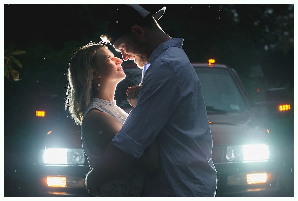 Couple embraces in front of a vehicle with headlights on, illuminated at night.