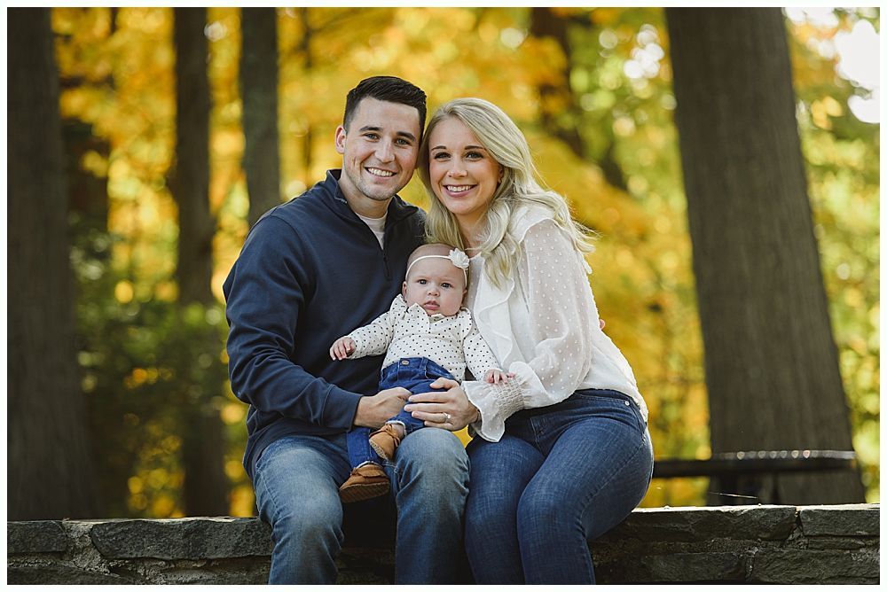 Family of three smiling outdoors, surrounded by fall foliage.