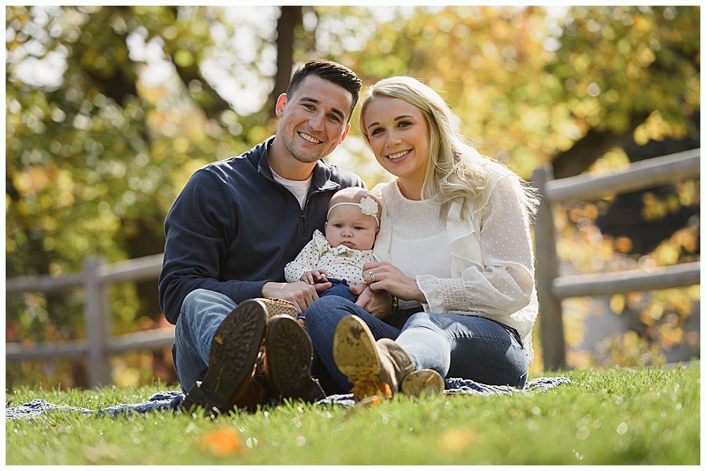 Family of three smiling outdoors, baby held on lap. Autumn backdrop of trees and fence.