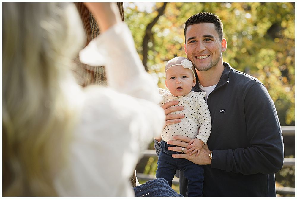 Father holding baby, both smiling. Blonde woman in foreground takes photo outdoors.